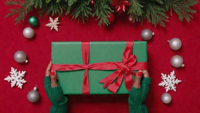 The primary subject of the image is a person holding a green and red gift-wrapped present. The person is wearing green sweaters and has red nail polish on their fingernails. The setting is a Christmas-themed background, featuring a tree and ornaments. The visual style is a photo, capturing the person's hand holding the present. The colors and mood of the image are festive and joyful, as it depicts a holiday celebration.